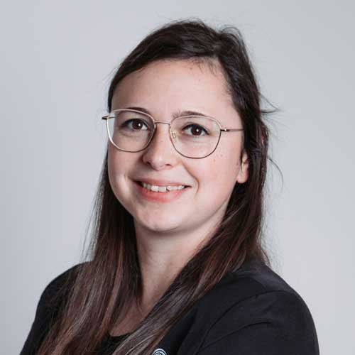 Porträt einer lächelnden jungen Frau mit langen dunklen Haaren, Brille und schwarzer Kleidung vor neutralem Hintergrund – professionelles Business-Profilfoto.