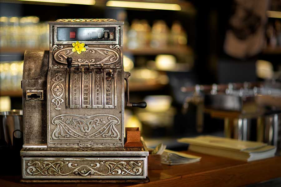 Registrierkassa_900x600 Antike Registrierkasse aus Metall mit floralen Verzierungen und gelber Blume auf der Vorderseite, aufgestellt auf einer hölzernen Theke in einem stimmungsvoll beleuchteten Café oder Restaurant.