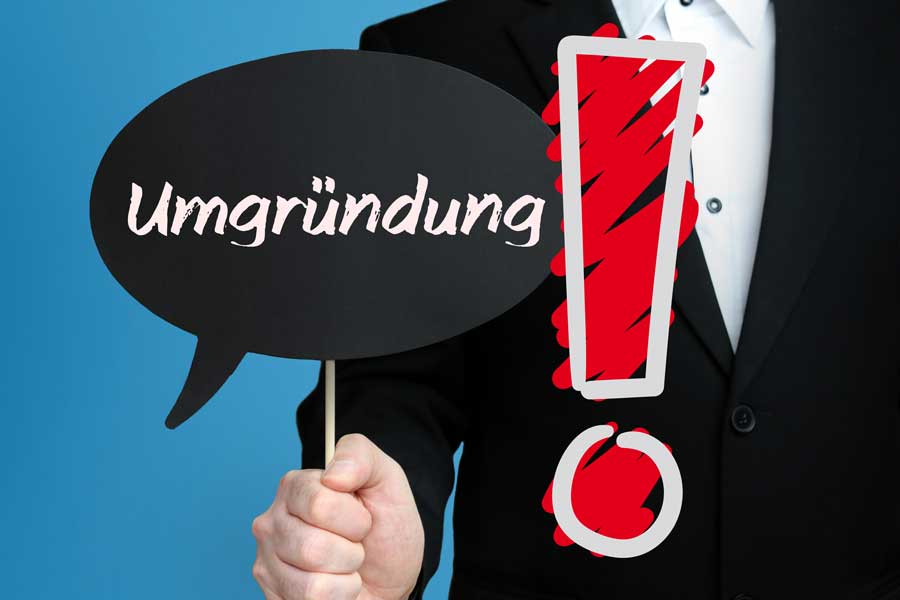 Umgruendung_900x600 Mann im Anzug hält eine Sprechblase mit dem Wort „Umgründung“ und ein großes rotes Ausrufezeichen – Symbolbild für Unternehmensumstrukturierung oder rechtliche Firmenänderung.