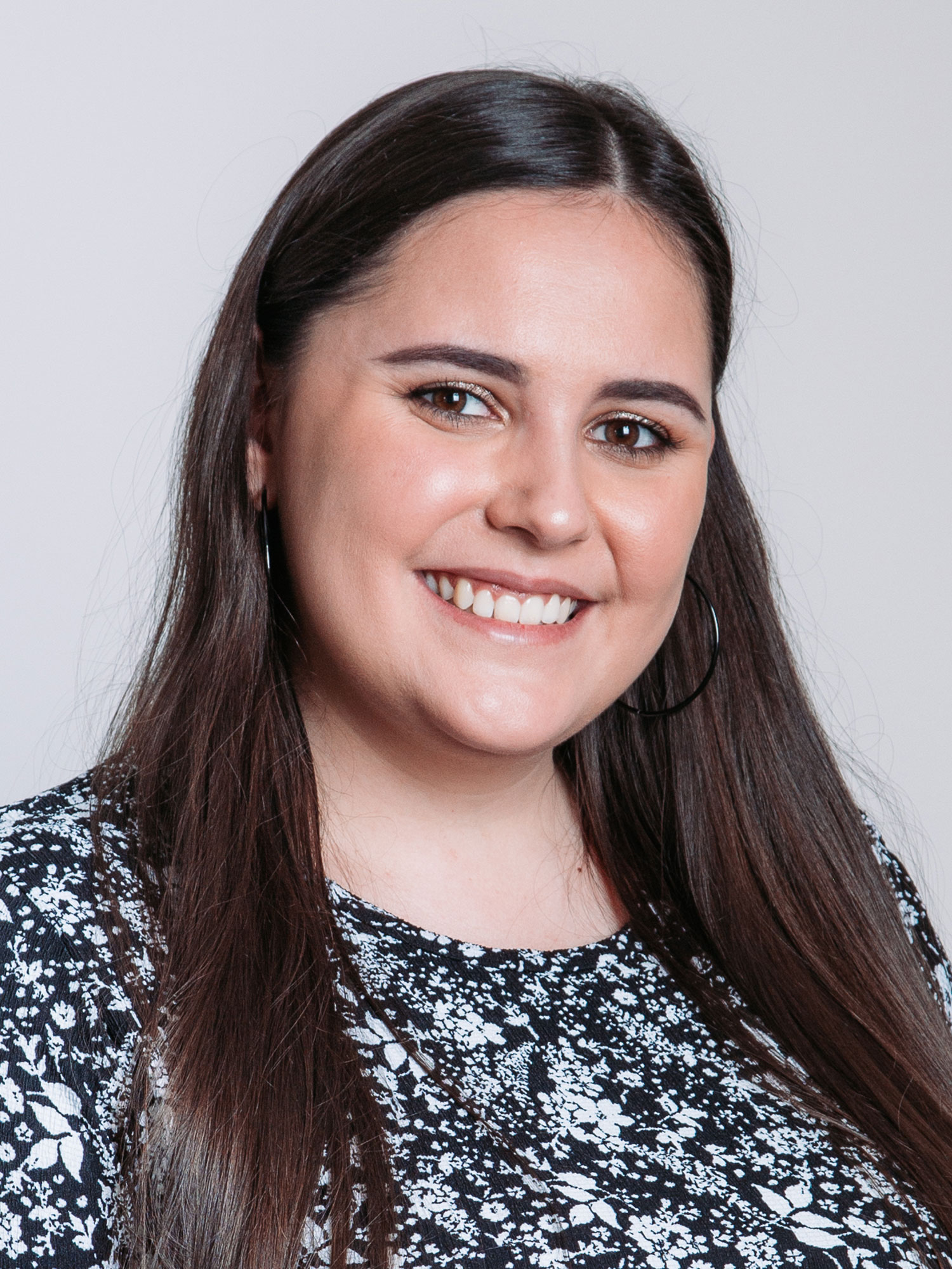 Claudia-S_1500x2000 Porträt einer jungen Frau mit langen, glatten braunen Haaren, die freundlich in die Kamera lächelt. Sie trägt große, runde Ohrringe und ein schwarzes Oberteil mit weißem Blumenmuster. Der Hintergrund ist neutral hellgrau