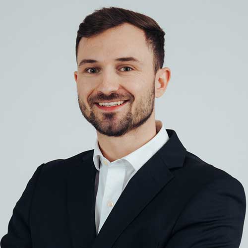 Porträt eines lächelnden jungen Mannes mit kurzem braunem Haar und Bart, der in einem dunklen Anzug und weißem Hemd vor neutralem Hintergrund steht – professionelles Businessfoto von Christoph K.