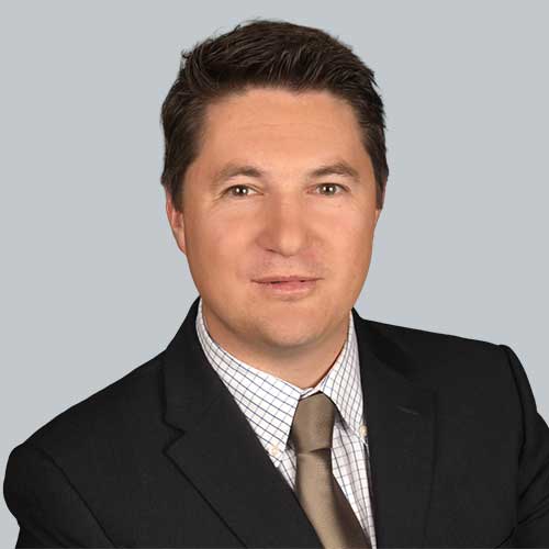 Porträtfoto von Michael Binder, einem Mann mit kurzen braunen Haaren, hellem Teint und braunen Augen, der freundlich in die Kamera blickt. Er trägt einen dunklen Anzug, ein weiß kariertes Hemd und eine beige-goldene Krawatte, vor einem neutral grauen Hintergrund.