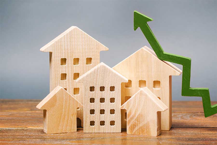 Holzmodelle von Wohnhäusern in verschiedenen Größen stehen nebeneinander auf einem Holztisch, während ein grüner, nach oben zeigender Pfeil im Hintergrund einen Anstieg symbolisiert – Darstellung steigender Mietpreise oder Immobilienwerte.