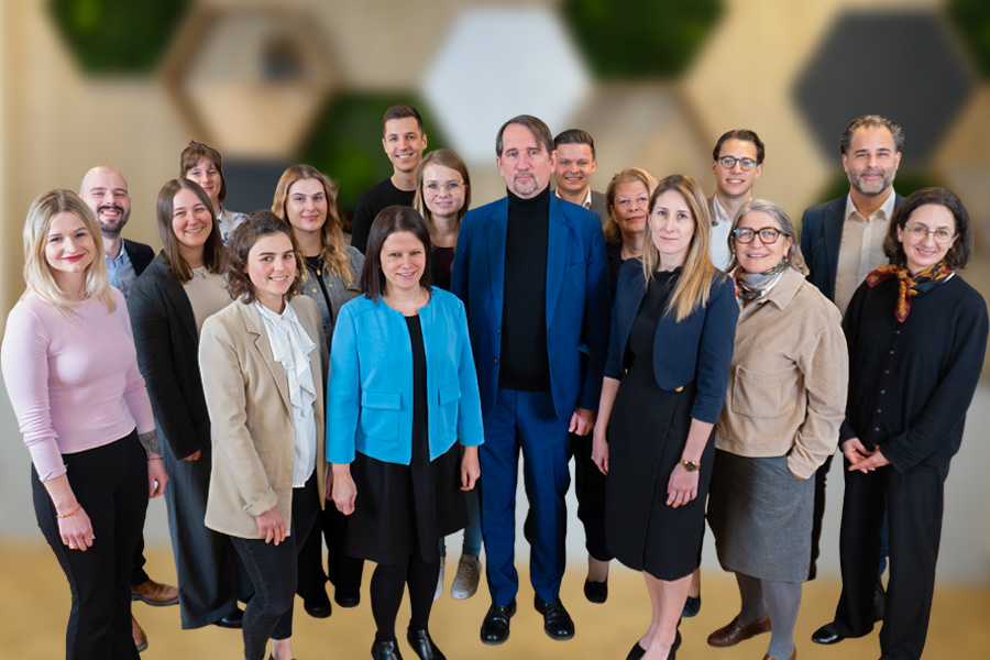 Gruppenfoto des Teams mit 15 Mitarbeitenden – Männer und Frauen unterschiedlichen Alters in Business-Kleidung, posierend in einem modernen Büro mit dekorativen Waben-Wandelementen und Begrünung im Hintergrund.