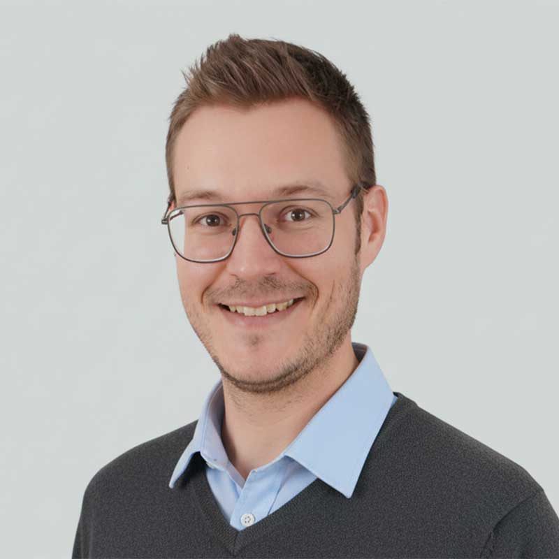 Porträt eines lächelnden Mannes mit kurzen, dunklen Haaren vor einem neutralen, hellen Hintergrund. Er trägt ein weißes Hemd und einen dunklen Anzug, was einen professionellen und gepflegten Eindruck vermittelt. Sein freundlicher Gesichtsausdruck strahlt Kompetenz und Offenheit aus.
