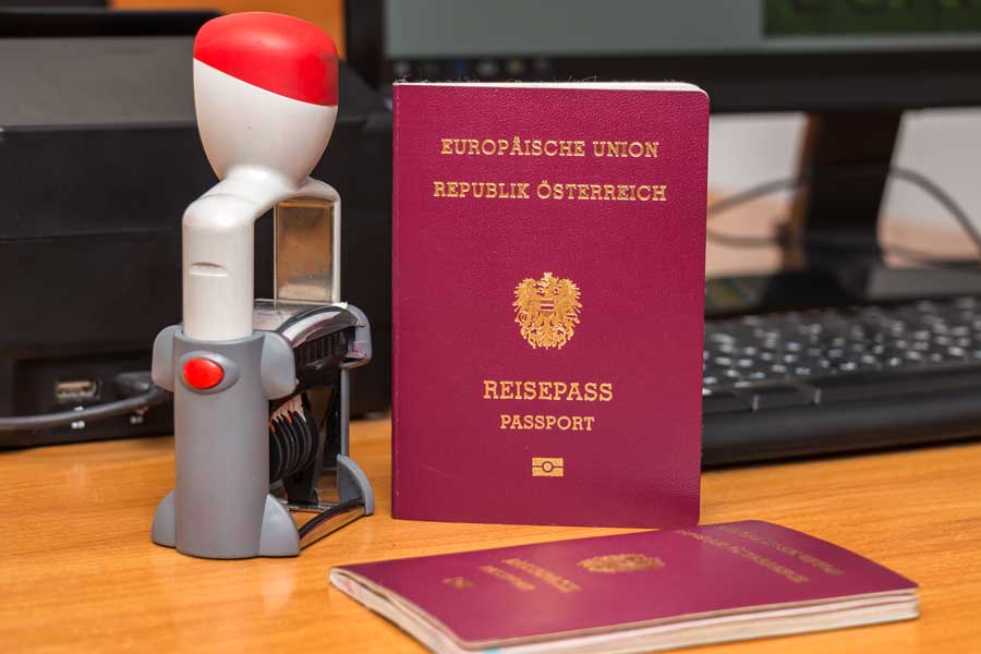 17:10 Österreichischer Reisepass der Europäischen Union auf einem Holzschreibtisch, daneben ein grauer Selbststempel mit roter Kappe und ein zweiter aufgeschlagener Reisepass, im Hintergrund ein schwarzer Computer-Monitor und eine Tastatur.