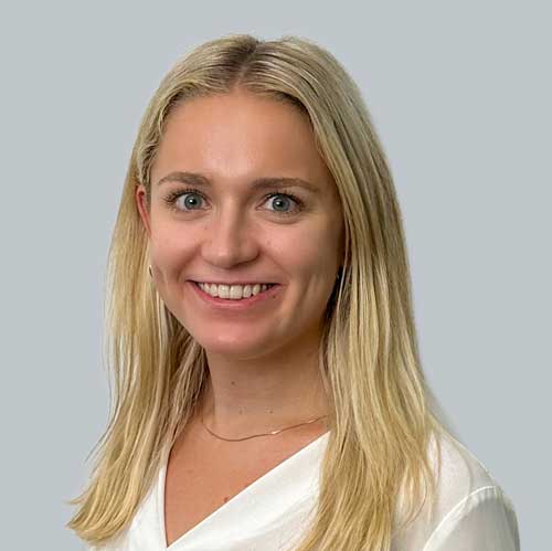 Porträtfoto von Elisabeth G. – junge Frau mit braunem, hochgestecktem Haar und Pony, die freundlich in die Kamera lächelt. Sie trägt eine weiße Bluse mit V-Ausschnitt und einen beigefarbenen Blazer vor einem hellgrauen Hintergrund.