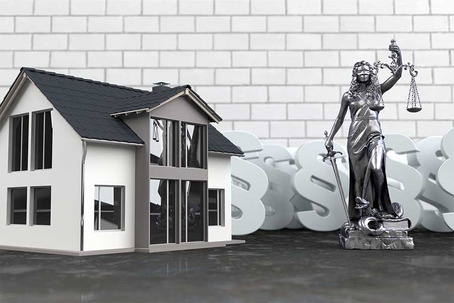 Modell eines Einfamilienhauses neben einer Justitia-Figur mit Waage und Schwert, im Hintergrund weiße Paragrafenzeichen vor einer Backsteinwand – Symbolbild für Immobilienrecht und gesetzliche Regelungen rund um Wohneigentum.