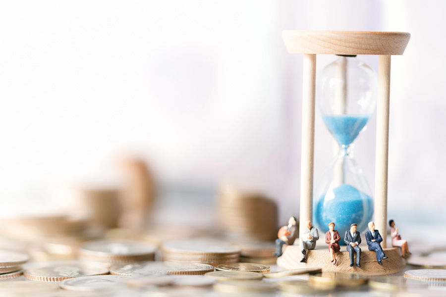 Miniaturfiguren von Senioren sitzen auf dem Sockel einer Sanduhr mit blauem Sand, die auf verstreuten Euro-Münzen steht – symbolisch für Altersvorsorge, Pension und den Zusammenhang von Zeit und Geldanlage.